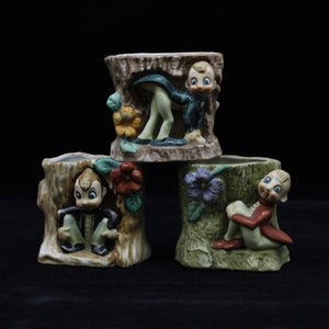 May include: Three ceramic planter pots shaped like tree stumps, each featuring a different whimsical elf figurine. The elf figurines are painted in bright colors, including green, red, blue, and yellow. The pots are perfect for adding a touch of whimsy to any home decor.