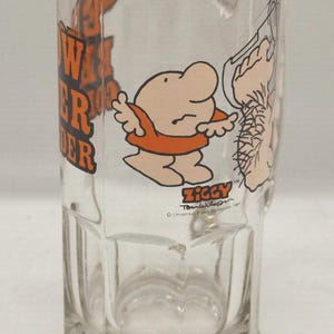 May include: Clear glass beer mug featuring a cartoon character named Ziggy in an orange shirt. The mug has the words "Power Wonder" in orange lettering and the Ziggy logo. The mug is a classic design with a rectangular base.