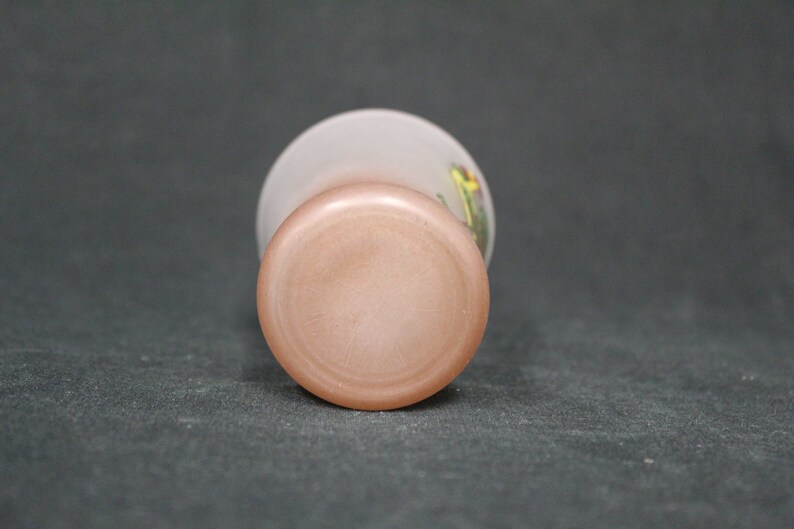 May include: The base of a frosted glass vase with a pink tint. The vase features a floral design with yellow, green, and red accents.