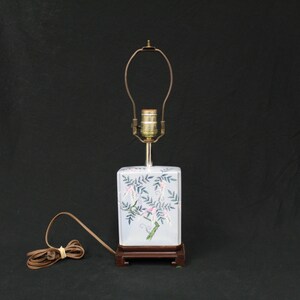 May include: A white ceramic table lamp with a square base featuring a floral design in shades of pink, green, and blue. The lamp has a brown wooden base and a simple metal shade.