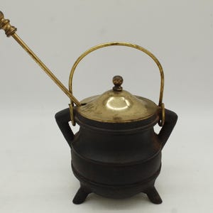 May include: A small, dark brown cast iron cauldron with a brass lid, handle, and stirrer. The cauldron has three small feet and two handles. The lid is topped with a small brass knob. The stirrer is long and thin, with a decorative brass ball at the end.