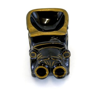 May include: Black ceramic ashtray shaped like a vintage car with gold accents. The car has two large headlights and a deep bowl for ashes.