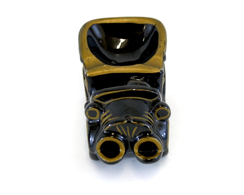 May include: Black ceramic ashtray shaped like a vintage car with gold accents. The car has two large headlights and a deep bowl for ashes.