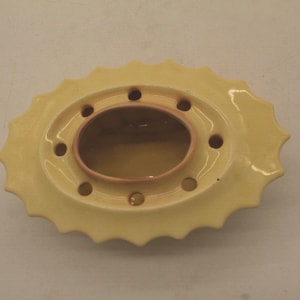 May include: A pale yellow ceramic flower frog with a scalloped edge and an oval center. The frog has multiple holes for arranging stems and a contrasting pinkish-brown interior.