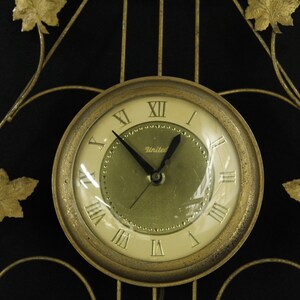 May include: A gold-toned wall clock with Roman numeral markings and a black face. The clock is set against a black background and features a decorative metal frame with gold leaf accents.