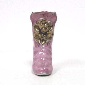 May include: A small, decorative ceramic boot in a soft pink hue. The boot features a gold-colored floral design on the front and is likely a collectible or decorative item. The boot's design is detailed and appears to be hand-painted.