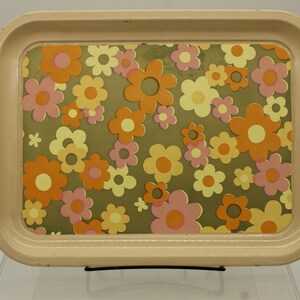 May include: A rectangular, cream-colored metal tray with rounded corners. The tray's interior features a floral pattern with orange, pink, and yellow flowers on a green background. The tray is supported by a black metal stand.