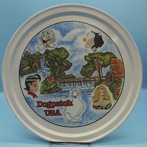 May include: Round metal tray with a white rim and a colorful illustration of Dogpatch USA. The central image depicts a landscape with waterfalls, a bridge, and cartoon characters. The text "Dogpatch USA" is prominently displayed.