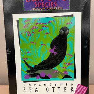 May include: A jigsaw puzzle box featuring an illustration of a sea otter. The box has the words "ENDANGERED SPECIES JIGSAW PUZZLES" at the top. The sea otter is black with purple feet and is surrounded by green seaweed and purple fish.
