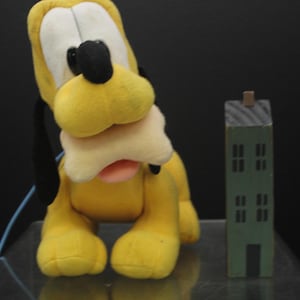 May include: A plush yellow Pluto dog toy with a cream-colored muzzle and black ears. A small, green wooden building stands beside the toy. The toy dog has a pink tongue and is sitting upright.