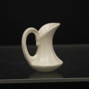 May include: A small, white ceramic pitcher with a curved spout and a handle. The pitcher is sitting on a black surface.