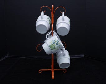 May include: A set of six white ceramic mugs with floral designs are displayed on a metal mug tree. The tree is orange and has a decorative swirl design.