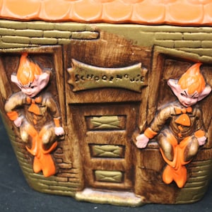 May include: Vintage ceramic cookie jar shaped like a schoolhouse. The jar features two elf-like figures with orange hair and clothing, flanking a door with the words "School House". The roof is orange, and the walls are brown and gold.