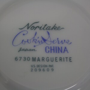 Puede incluir: Primer plano de un plato de cer&aacute;mica blanca con el texto "Noritake Cookin' Serve" en escritura verde y azul. El plato est&aacute; marcado "Japan CHINA" y "6730 MARGUERITE". Tambi&eacute;n tiene impreso "U.S. DESIGN PAT. 209609".