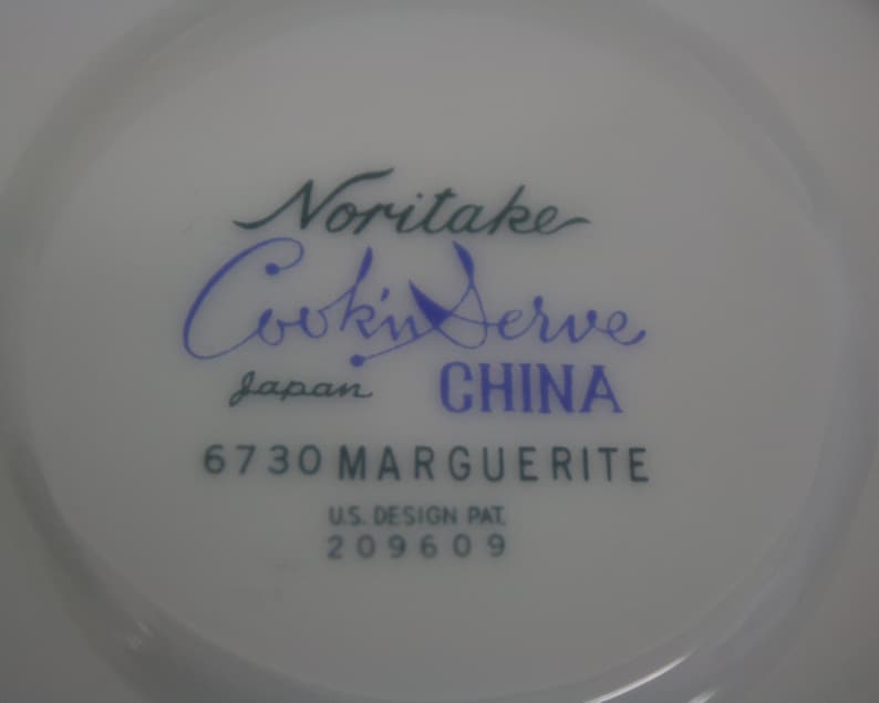 Puede incluir: Primer plano de un plato de cer&aacute;mica blanca con el texto "Noritake Cookin' Serve" en escritura verde y azul. El plato est&aacute; marcado "Japan CHINA" y "6730 MARGUERITE". Tambi&eacute;n tiene impreso "U.S. DESIGN PAT. 209609".