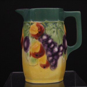 May include: A yellow ceramic pitcher with a green top and a handle. The pitcher is decorated with a raised design of grapes and peaches.