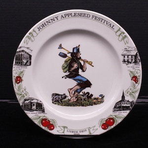 May include: White ceramic plate with a colorful illustration of Johnny Appleseed holding a shovel and walking through a field. The plate has a blue and white border with a red apple design. The text "Johnny Appleseed Festival" is written in a black cursive font around the top edge of the plate. The text "Lisbon, Ohio 1999" is written in black at the bottom of the plate.