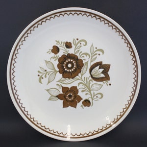 May include: A white ceramic dinner plate with a brown and green floral design. The plate has a scalloped edge.