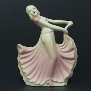 May include: A porcelain figurine of a woman in a flowing pink dress, holding the fabric. The figurine has blonde hair, red lips, and is posed in a graceful dance. The dress has a light pink skirt and a white bodice.