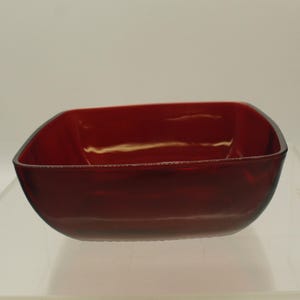 May include: A square, red glass bowl with a smooth, glossy finish. The bowl is empty and sits on a white surface.