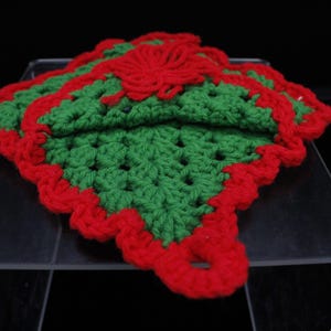 May include: A green and red crocheted pot holder with a scalloped edge. The pot holder is made of yarn and has a floral design in the center.