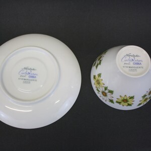 Puede incluir: Juego de taza y platillo blancos con un estampado floral de margaritas. La taza tiene un asa y el platillo es redondo. El juego est&aacute; marcado con "Noritake Cook & Serve China" y "6730 MARGUERITE".