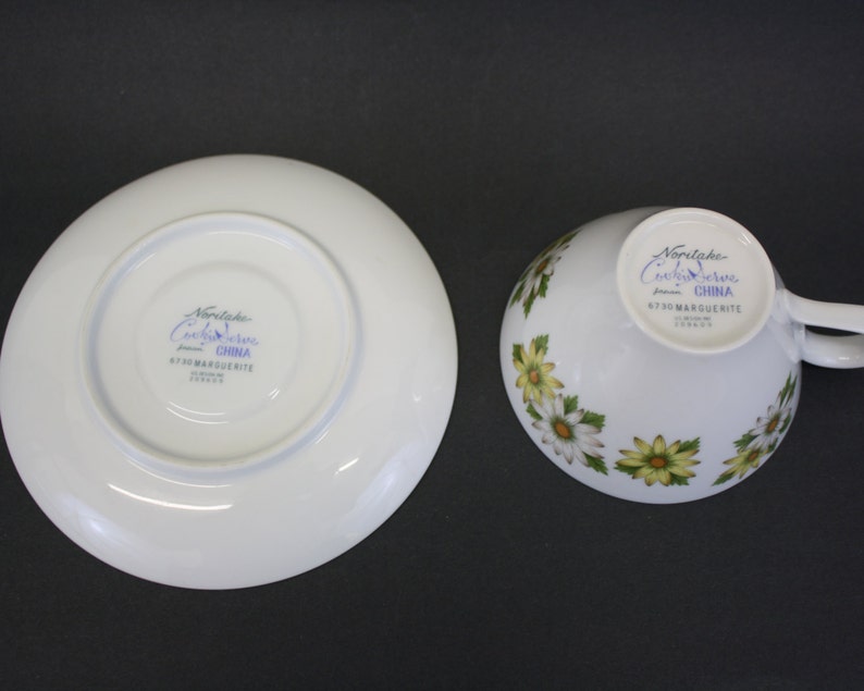 Puede incluir: Juego de taza y platillo blancos con un estampado floral de margaritas. La taza tiene un asa y el platillo es redondo. El juego est&aacute; marcado con "Noritake Cook & Serve China" y "6730 MARGUERITE".