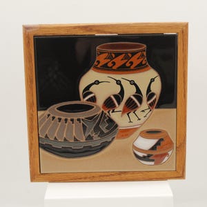 May include: A framed ceramic tile featuring three decorative pottery vessels. The largest vase is cream-colored with black bird illustrations and orange accents. The other two vessels are brown and tan with geometric designs.