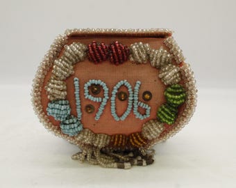 May include: A small, antique, peach-colored beaded box. The box is decorated with light blue beads spelling out "1906" and is framed by a beaded border. The box has a beaded fringe at the bottom and red and green beaded accents.