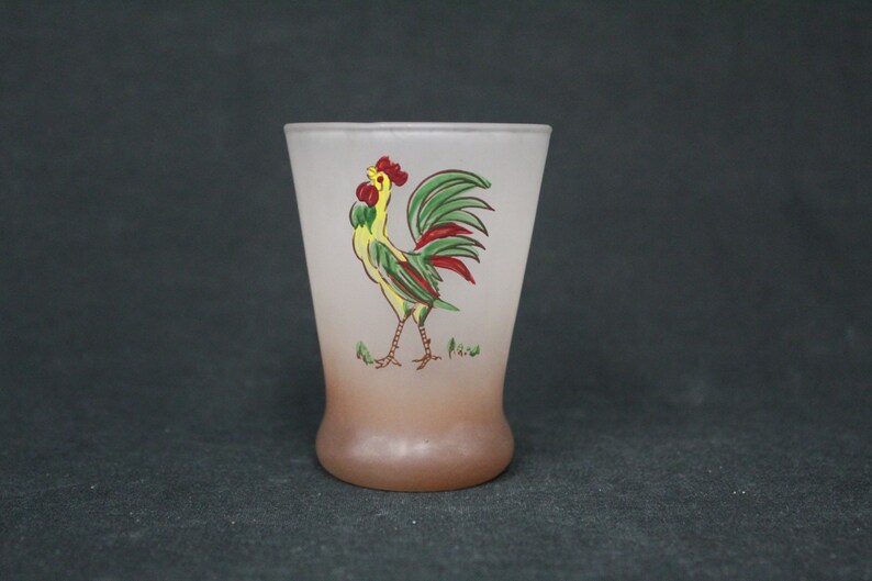 May include: A frosted glass shot glass featuring a rooster design. The rooster is yellow, green and red. The glass has a brown ombre effect at the bottom.