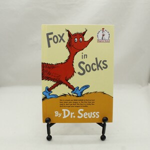 May include: A yellow and orange book cover with a red fox wearing blue socks. The book title is "Fox in Socks" by Dr. Seuss. The book is part of the "I Can Read It All By Myself" Beginner Books series.