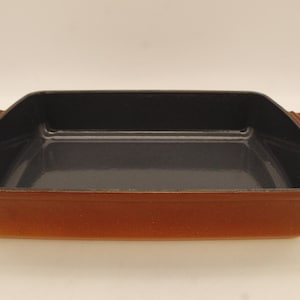 May include: A rectangular, brown cast iron baking dish with a dark interior. The dish has a slightly flared rim and two handles on either side. The interior is a dark gray color.