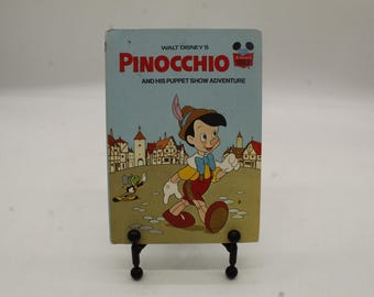May include: A vintage Walt Disney's Pinocchio book. The cover features Pinocchio in a red outfit with a yellow shirt and a blue bow tie. The title is in red and the background is a light blue color.
