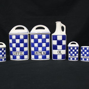 May include: A set of seven ceramic kitchen canisters and containers. The pieces are white with a blue checkered pattern. The containers are labeled "Cinnamon", "Coffee", "Oatmeal", "Oil", "Pepper", and "Cloves".