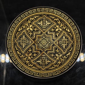 May include: A round, decorative object with an intricate design. The object features a black background with gold-colored, ornate patterns. The design includes floral and geometric elements, with a border of repeating shapes.