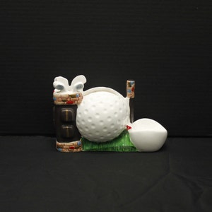 May include: Ceramic golf-themed desk organizer with a golf bag, golf ball, and a small dish. The golf bag is brown with red, white, and blue accents. The golf ball is white with dimples. The dish is white.