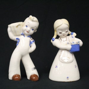 May include: A pair of vintage ceramic figurines, a boy carrying a bucket and a girl holding a blue basket. Both figurines are painted in white with blue and brown accents.