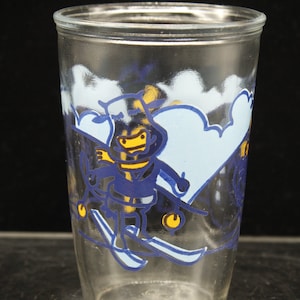 May include: A clear glass drinking glass with a blue and yellow cartoon illustration of a person skiing down a snowy mountain. The person is wearing a blue hat, scarf, and jacket. The background is blue and white.