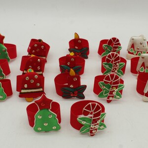 May include: A collection of festive Christmas napkin rings. Each ring is red and adorned with various holiday-themed decorations, including Christmas trees, candles, candy canes, and houses. The decorations feature green, white, and gold accents.