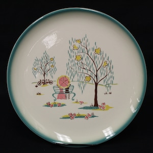 May include: A white ceramic dinner plate with a teal rim features a whimsical scene of two figures sitting on a bench under a weeping willow tree. The figures are holding an umbrella and are surrounded by yellow flowers.