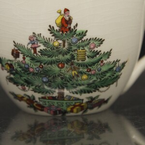 May include: A white ceramic teacup with a Christmas tree design. The tree is decorated with ornaments, lights, and presents. A small figure of a man with a sack on his back stands at the top of the tree.