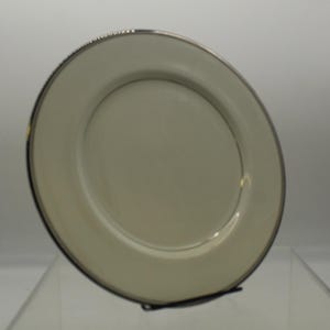 May include: A white ceramic plate with a thin black and gold rim. The plate is slightly angled and appears to be new.