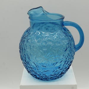 May include: A blue glass pitcher with a textured surface and a handle. The pitcher has a round body and a spout for pouring. The pitcher is sitting on a white surface.