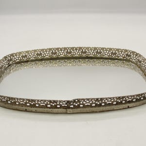 May include: Rectangular silver-toned vanity tray with a mirrored surface. The tray's border features a delicate, openwork design. The mirrored surface reflects the surrounding environment. A decorative item for displaying jewelry or perfumes.