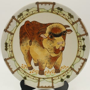 May include: A white ceramic plate with a brown and yellow bull illustration in the center. The plate has a green and brown border with a repeating pattern of fences.