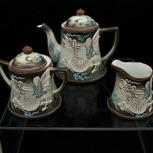 May include: A set of three ceramic teacups and teapot decorated with a dragon design. The teapot is brown with white and blue accents. The teacups are white with brown and blue accents.