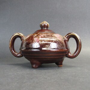 May include: A brown ceramic sugar bowl with a lid and two handles. The bowl has a glossy finish with reddish-pink accents and sits on three small feet. The lid is topped with a small knob.