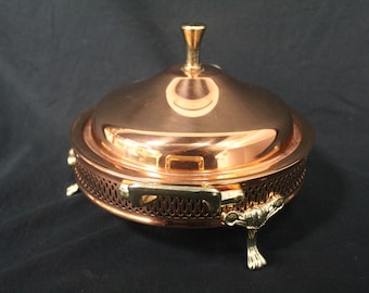 May include: A copper chafing dish with a brass lid and three decorative brass feet. The dish has a decorative, woven pattern around the base.