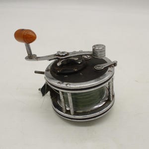 May include: A vintage fishing reel with a silver frame, black top, and a wooden handle. The reel has a spool of green fishing line and is designed for saltwater fishing. The reel is a classic piece of fishing equipment.