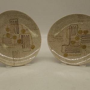 May include: Two white ceramic plates with a brown and gold geometric design. The plates have a speckled texture.